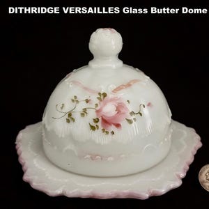 May include: White glass butter dome with a matching plate. The dome is decorated with a pink rose and green leaves. The plate and the dome have a scalloped edge with pink trim. The text "DITHRIDGE VERSAILLES Glass Butter Dome" is at the top.