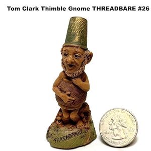 May include: A ceramic gnome figurine with a thimble hat, holding a basket. The gnome has a detailed face and beard. The base is green with the text "THREADBARE" and "1987". A quarter coin is next to the figurine for size comparison. The text "Tom Clark Thimble Gnome THREADBARE #26" is at the top.