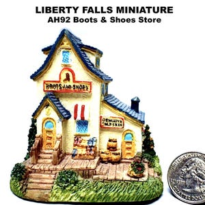 May include: A miniature Boots & Shoes store with a cream-colored facade, blue roof, and a small porch. The sign reads "Boots and Shoes" and "Genuine Calfskin". A quarter is next to the miniature for scale. The store is set on a green base.
