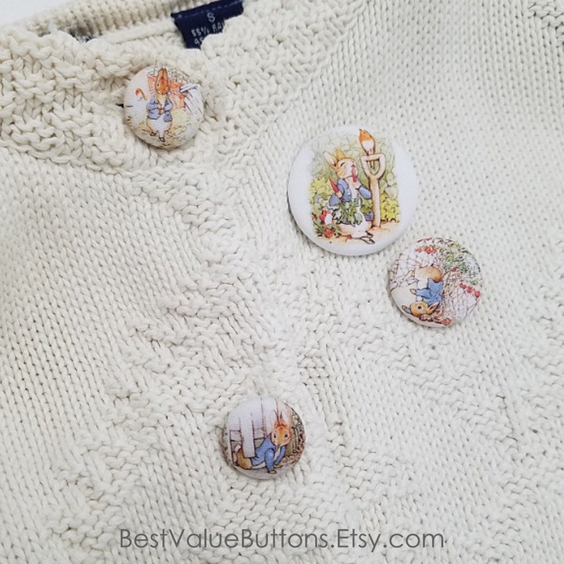 May include: Four white round buttons with images of Peter Rabbit from the children's book series by Beatrix Potter. The buttons are on a white knitted sweater.