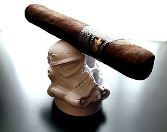 May include: A brown cigar rests on a white Stormtrooper helmet-shaped cigar holder. The cigar has a dark band with gold and red accents. The holder is on a black surface.