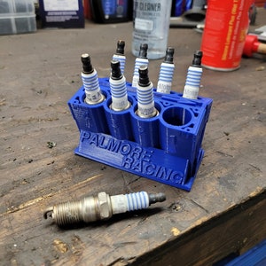 May include: A blue 3D-printed spark plug holder with the text "PALMORE RACING" holds several spark plugs. One spark plug rests on the wooden surface. The spark plugs have a silver body, black top, and white and blue insulator.