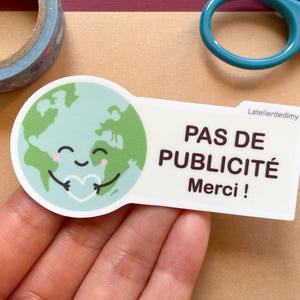 Peut inclure: Un autocollant avec une illustration de la Terre en dessin animé tenant un cœur. Le texte sur l'autocollant est "Pas de publicité Merci !". L'autocollant est blanc avec du texte noir et se trouve sur une surface brune.
