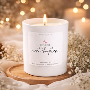 Könnte beinhalten: Eine weiße Kerze mit brennendem Docht, mit dem Text "Here's to your next chapter" auf einem Etikett. Die Kerze steht auf einem Holzuntersetzer, umgeben von Blumen und Bokeh-Lichtern. Das Etikett gibt auch "100% Vegan Soy Wax" und "Brenndauer: 25 Stunden" an.
