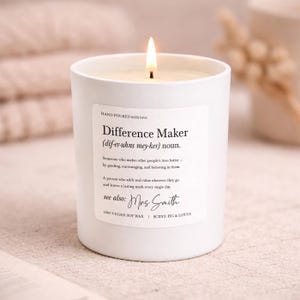 Puede incluir: Una vela blanca encendida con la inscripción "Difference Maker" en la etiqueta. La vela está en un recipiente blanco y tiene una mecha encendida. La etiqueta también incluye el texto "HAND POURED with love" y "100% VEGAN SOY WAX".