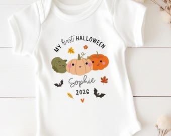 Puede incluir: Un body blanco de manga corta para bebé con un gráfico de tres calabazas sonrientes en verde, naranja claro y naranja. El texto dice "MY first HALLOWEEN", "Sophie" y "2026", acompañado de pequeñas ilustraciones de murciélagos, hojas y un corazón.