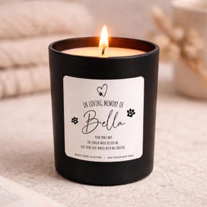 Puede incluir: Una vela negra encendida con una etiqueta blanca que dice "In Loving Memory of Bella". La etiqueta incluye huellas de patas y un corazón. El texto de abajo dice "Your paws mat, no longer walk beside me, but your love walks with me forever." La vela tiene un tiempo de combustión de 25 horas.