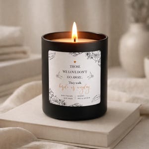 May include: A lit black candle with a white label featuring floral designs and the text "THOSE WE LOVE DON'T GO AWAY... They walk beside us everyday." The candle is on a stack of books.