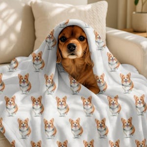 May include: A Cavalier King Charles Spaniel dog peeks out from under a white blanket. The blanket is covered in a repeating pattern of watercolor-style Corgi dogs. The dog has brown fur and is sitting on a white couch.