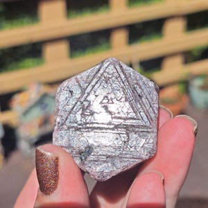 May include: A hexagonal, metallic-toned mineral specimen featuring a distinct triangular geometric pattern etched into its surface. The crystal displays a reflective, silvery-purple hue with visible striations and a small dark inclusion near the base.