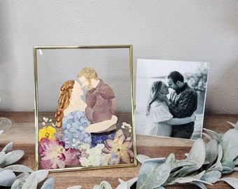May include: A framed pressed flower portrait of a couple embracing. The portrait is made with dried flowers and is displayed in a gold frame.