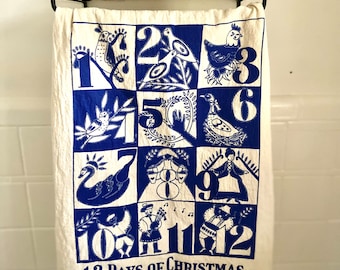 May include: A white kitchen towel with a blue design of the 12 Days of Christmas. Each day is represented by a number and a corresponding illustration. The text "12 Days of Christmas" is printed at the bottom of the towel.