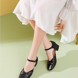 May include: Black leather ankle strap heeled shoes with cutouts and a buckle closure. The shoes have a low block heel.