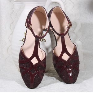 May include: A pair of burgundy glitter T-strap sandals with a delicate ankle strap and a small buckle fastening.