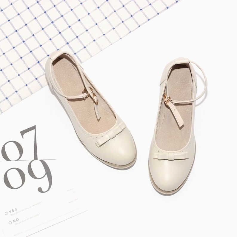 May include: A pair of cream-colored leather shoes with a bow detail and an ankle strap. The shoes have a closed toe and a low heel.