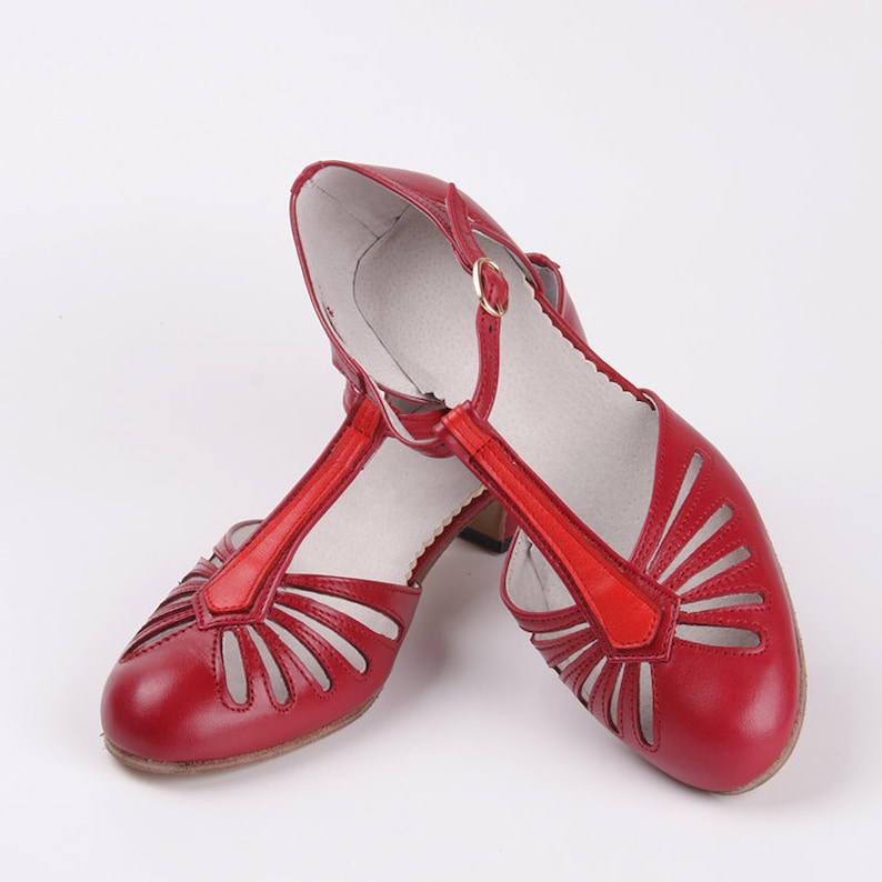 Pu&ograve; includere: Un paio di scarpe con tacco in pelle rossa con punta aperta e design a T-strap. Le scarpe hanno un motivo a taglio sul collo del piede e sui lati.