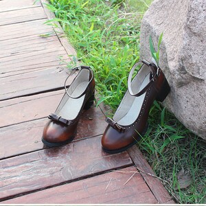May include: A pair of brown leather ankle strap shoes with a bow detail on the toe. The shoes are on a wooden surface with green grass in the background.