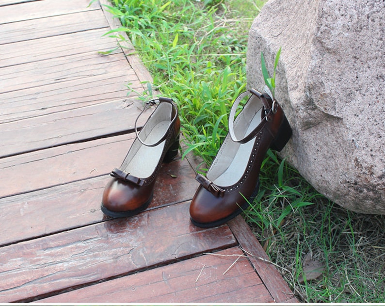 May include: A pair of brown leather ankle strap shoes with a bow detail on the toe. The shoes are on a wooden surface with green grass in the background.