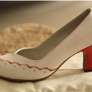 Pu&ograve; includere: Scarpa bianca con tacco alto con un bordo rosso attorno al bordo della punta e un tacco rosso.