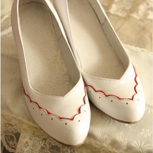 Pu&ograve; includere: Un paio di ballerine in pelle bianca con bordo rosso a festoni. Le scarpe hanno punta a punta e un design semplice.