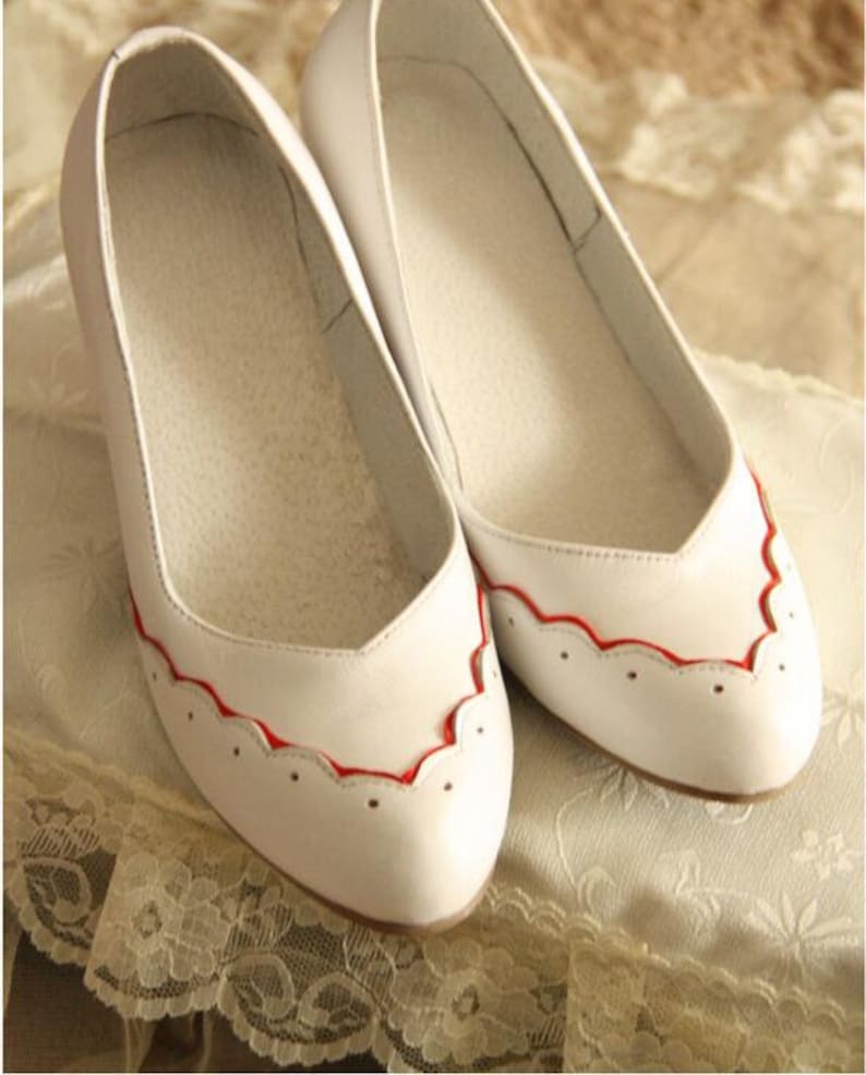 Pu&ograve; includere: Un paio di ballerine in pelle bianca con bordo rosso a festoni. Le scarpe hanno punta a punta e un design semplice.