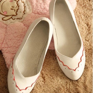 Pu&ograve; includere: Un paio di ballerine in pelle bianca con bordo rosso a festoni. Le scarpe sono su una superficie soffice di colore marrone chiaro.