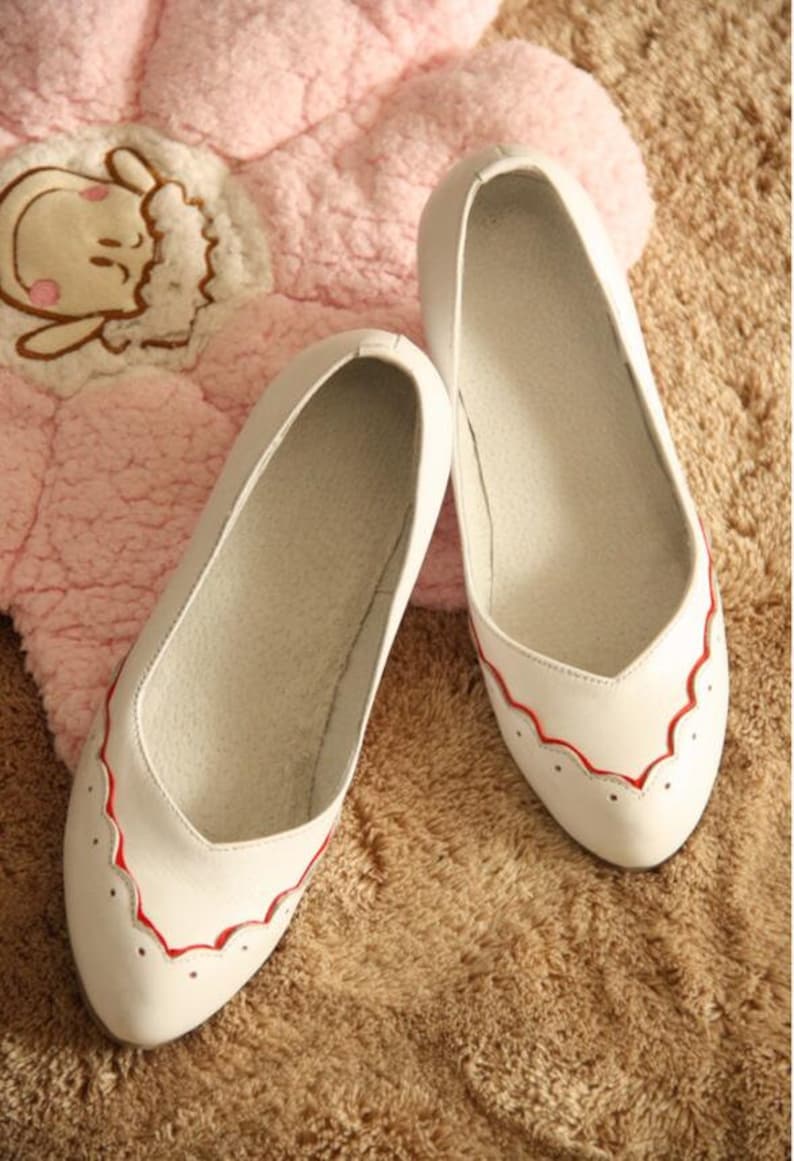 Pu&ograve; includere: Un paio di ballerine in pelle bianca con bordo rosso a festoni. Le scarpe sono su una superficie soffice di colore marrone chiaro.