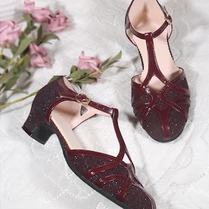 May include: A pair of burgundy heeled shoes with a T-strap and a sparkly finish. The shoes have a low heel and a closed toe.