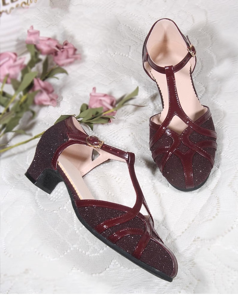 May include: A pair of burgundy heeled shoes with a T-strap and a sparkly finish. The shoes have a low heel and a closed toe.