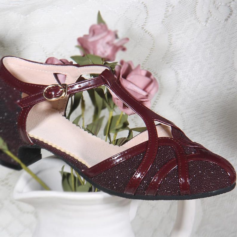 May include: Burgundy coloured women's dress shoe with a T-strap and a low heel. The shoe has a sparkly finish and a buckle closure.