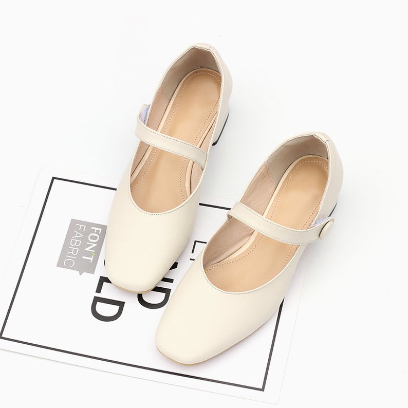 K&ouml;nnte beinhalten: Ein Paar cremefarbene Lederschuhe mit einem Riemen und einer Schnalle. Die Schuhe haben eine quadratische Spitze und einen niedrigen Absatz.