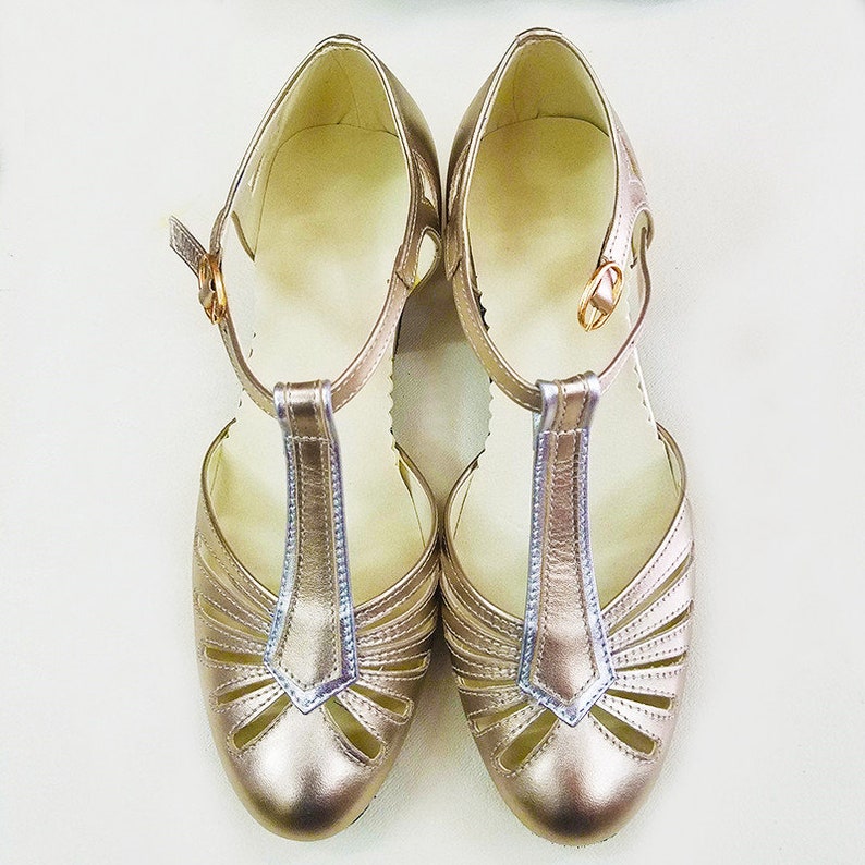 Pu&ograve; includere: Un paio di scarpe da donna color oro con un design a cinturino a T e dettagli traforati. Le scarpe hanno la punta chiusa e un tacco basso.