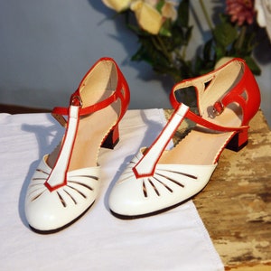 Pu&ograve; includere: Un paio di scarpe in pelle bianca con finiture rosse e tagli. Le scarpe hanno un design a cinturino a T e un tacco basso.