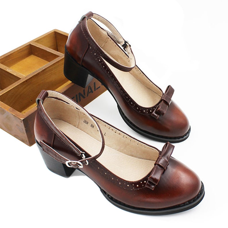 May include: A pair of brown leather ankle strap shoes with a bow detail on the toe. The shoes have a block heel and a closed toe.