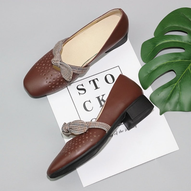 Puede incluir: Un par de zapatos de cuero marr&oacute;n con un lazo de tela a cuadros y un tac&oacute;n bajo de bloque. Los zapatos tienen un dise&ntilde;o perforado y est&aacute;n sobre una superficie blanca con la palabra "STOCK" impresa.