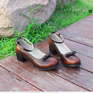 May include: A pair of brown leather ankle strap shoes with a bow detail on the toe. The shoes have a low block heel and are sitting on a wooden surface.