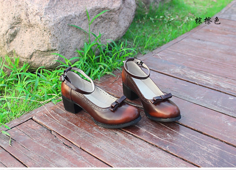 May include: A pair of brown leather ankle strap shoes with a bow detail on the toe. The shoes have a low block heel and are sitting on a wooden surface.