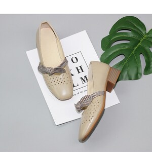 Puede incluir: Un par de zapatos de cuero beige con un lazo a cuadros y un peque&ntilde;o tac&oacute;n. Los zapatos tienen un dise&ntilde;o recortado y est&aacute;n sobre una superficie blanca.