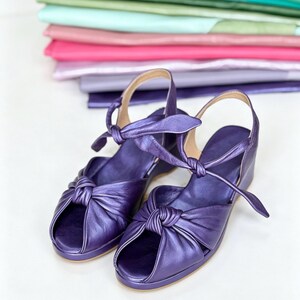 Op de afbeelding: Een paar metallic paarse leren sandalen met een open teen, een geknoopt detail aan de voorzijde en een verstelbare enkelband met striksluiting. De sandalen hebben een lage sleehak en een glad, effen voetbed.