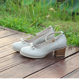 May include: A pair of white leather heeled shoes with a bow detail and ankle strap. The shoes are on a wooden surface.