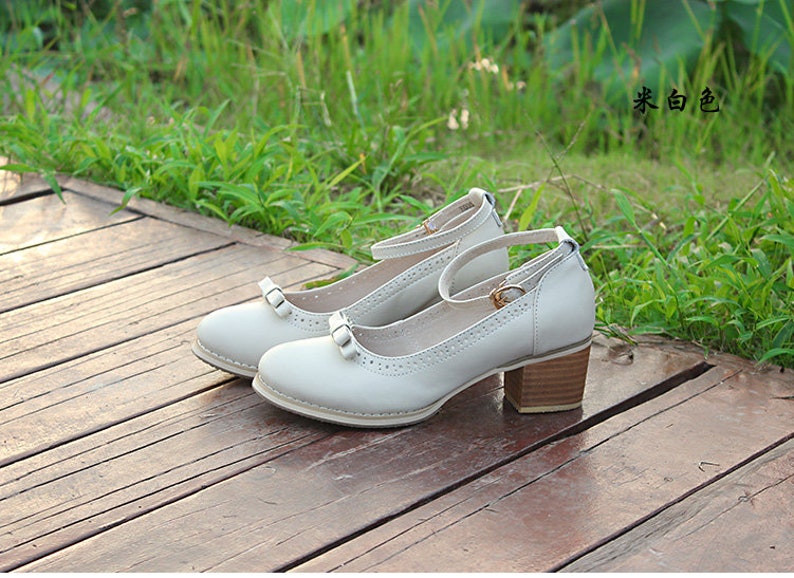 May include: A pair of white leather heeled shoes with a bow detail and ankle strap. The shoes are on a wooden surface.