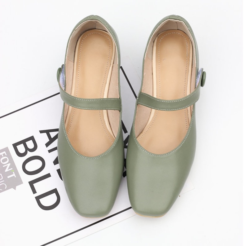 K&ouml;nnte beinhalten: Ein Paar gr&uuml;ne Lederschuhe mit Riemen und Schnalle. Die Schuhe haben eine quadratische Spitze und einen niedrigen Absatz.