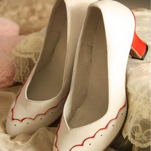 Pu&ograve; includere: Un paio di d&eacute;collet&eacute; in pelle bianca con bordi rossi. Le scarpe hanno un design a festoni e un piccolo tacco.
