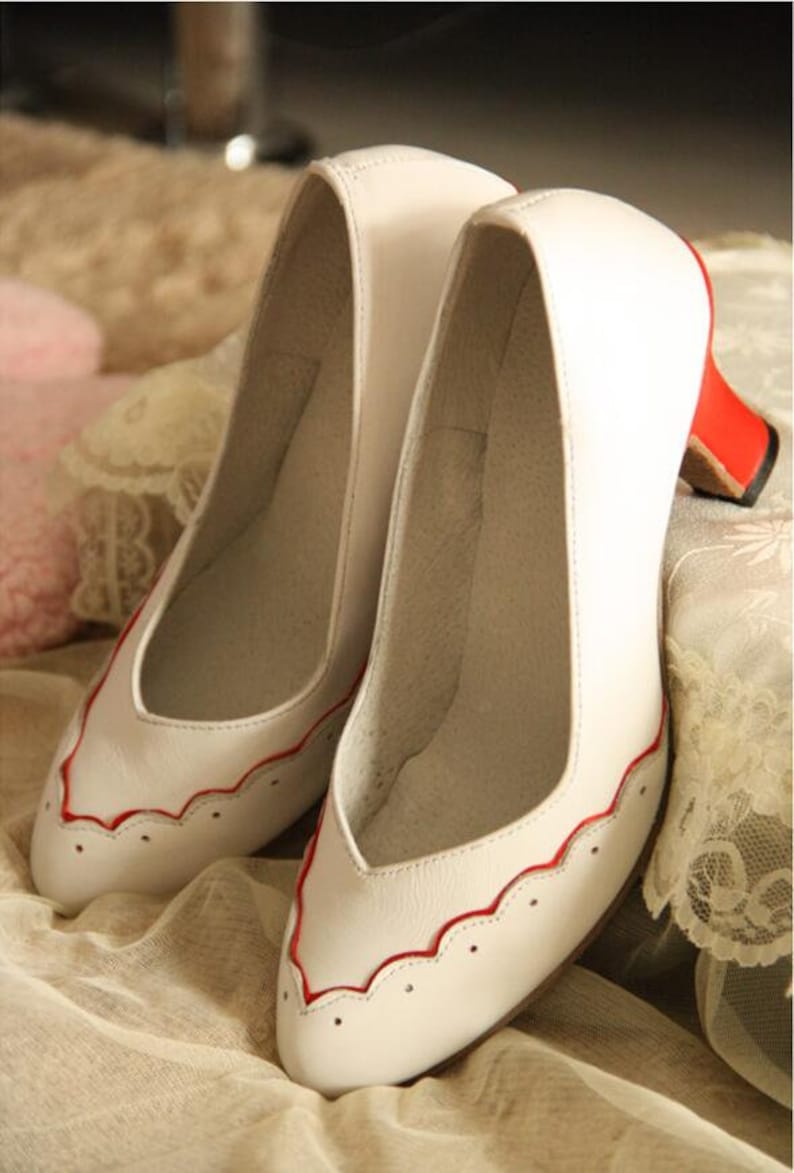 Pu&ograve; includere: Un paio di d&eacute;collet&eacute; in pelle bianca con bordi rossi. Le scarpe hanno un design a festoni e un piccolo tacco.