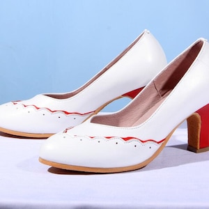 Pu&ograve; includere: Pumps in pelle bianca con bordo a festoni rosso e tacco rosso.
