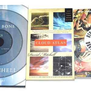 May include: Three hardcover books by David Mitchell are displayed. "The Bone Clocks" has a blue cover with concentric circles. "Cloud Atlas" features a collage of images. "Utopia Avenue" has a colorful, illustrated cover. All books are novels.