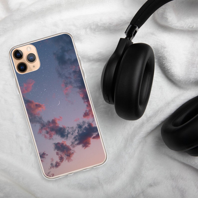 May include: A clear phone case with a pink and blue sunset sky and stars design. The case is on a white fuzzy surface with black headphones.