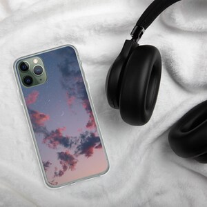 May include: A clear phone case with a pink and purple sunset sky and stars design. The case is on a white fuzzy surface next to black headphones.