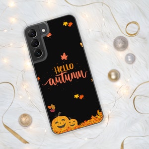 May include: A black phone case with a fall theme. The case features the words "Hello Autumn" in orange script, along with orange and yellow leaves and two jack-o'-lanterns. The case is on a white, fluffy surface.