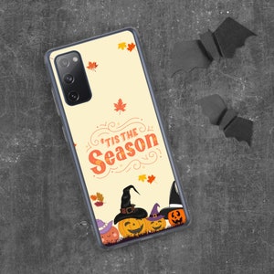 May include: A phone case with a Halloween theme. The case features the text "'Tis the Season" in orange and white, with illustrations of pumpkins wearing witch hats. Autumn leaves are scattered across the light yellow background.